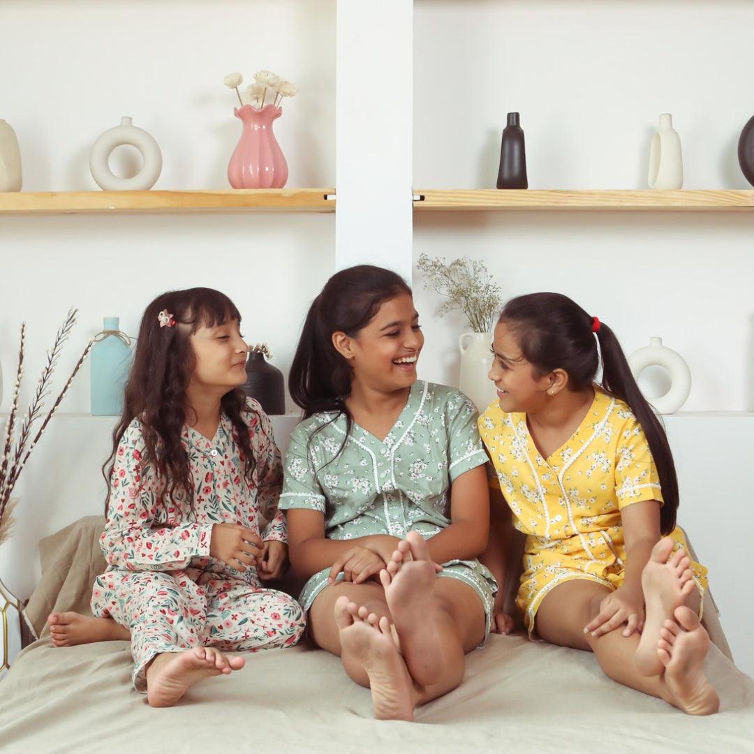 Three girls in colorful floral night suits sitting and smiling on a bed in a cozy room.