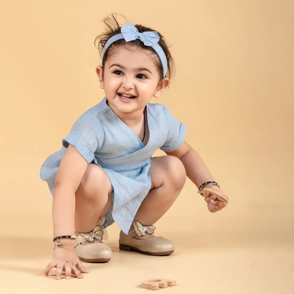 Smiling baby girl in a light blue dress and matching headband, squatting on a beige background