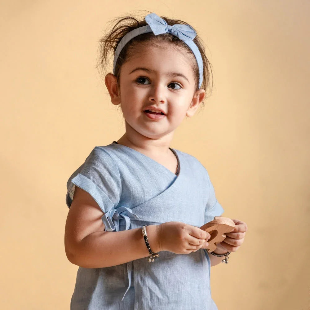 Smiling toddler girl in a light blue wrap dress and matching headband, holding a toy