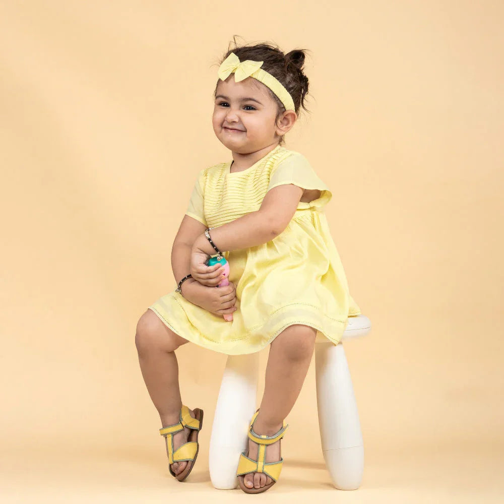 Smiling baby girl in yellow dress and headband sitting on stool, stylish kidswear outfit