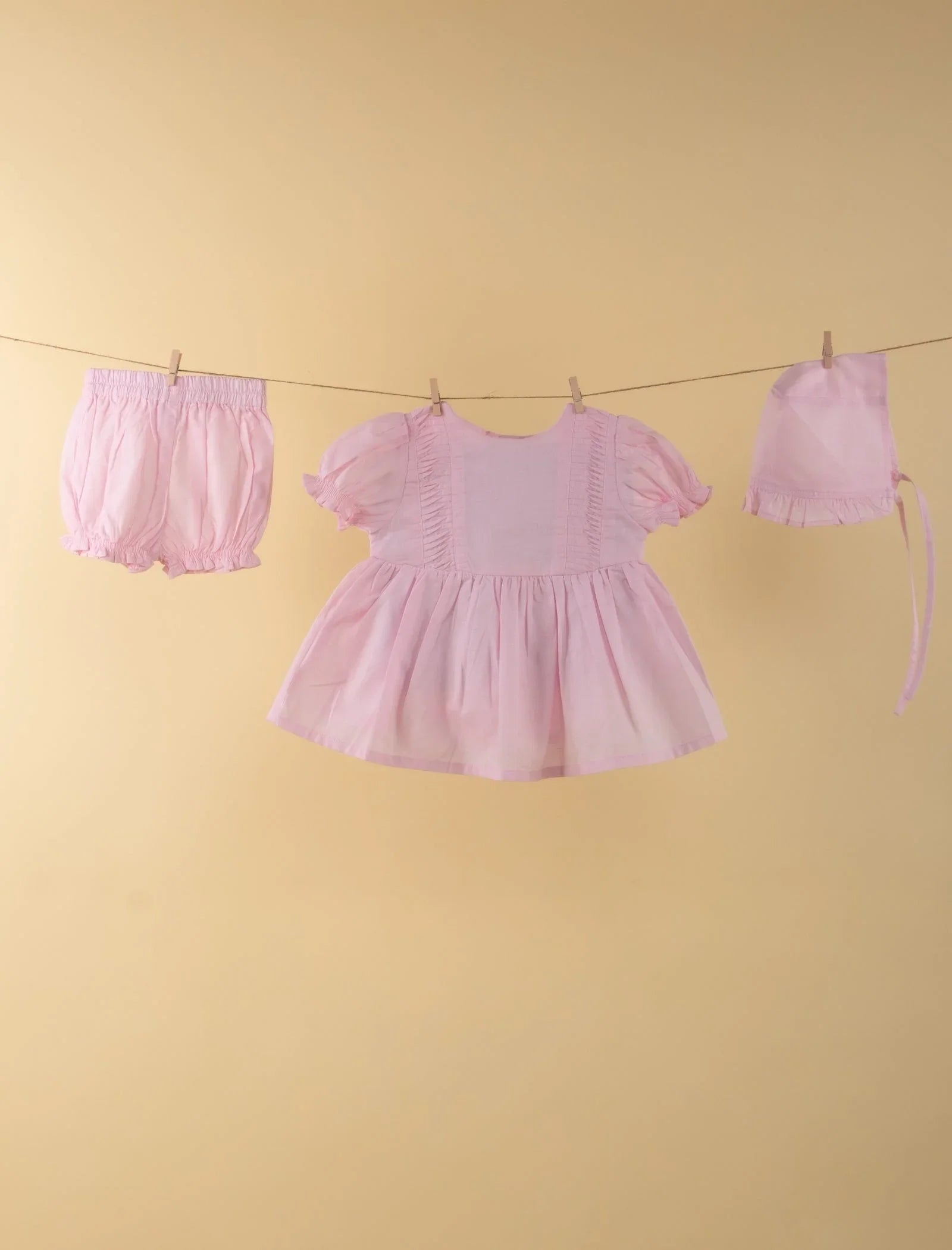 Light pink baby girl dress set with matching bloomers and bonnet hanging on a clothesline