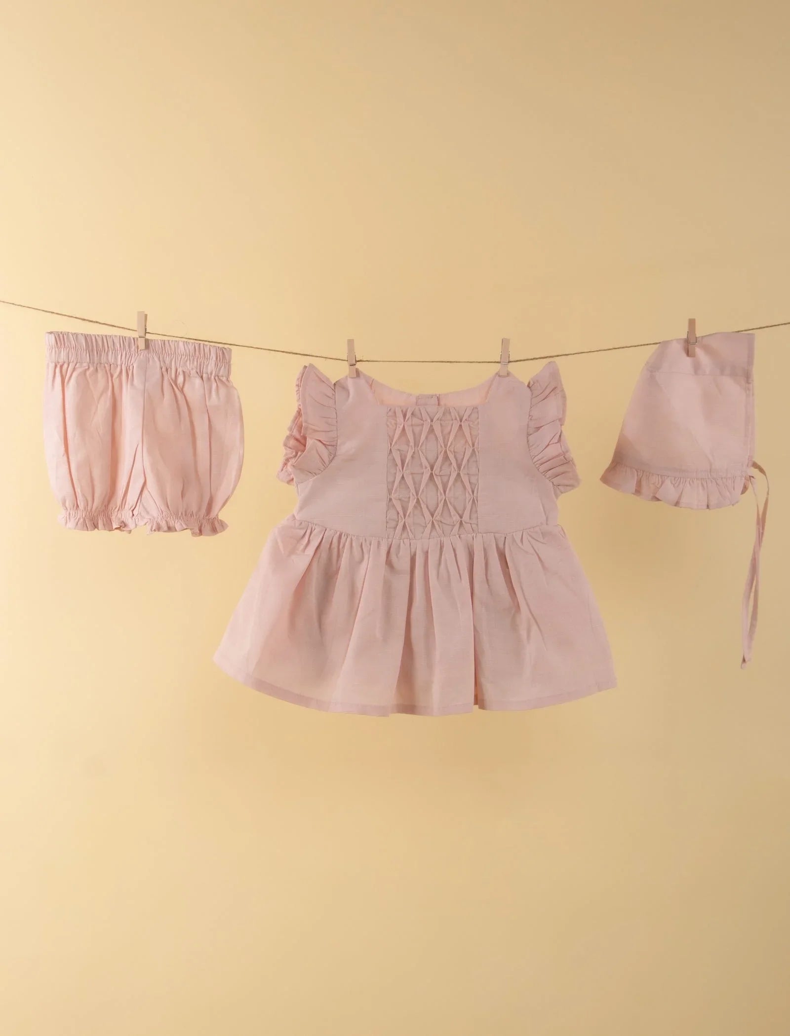 Light pink baby girl dress set with ruffled sleeves, bloomers, and bonnet on beige background