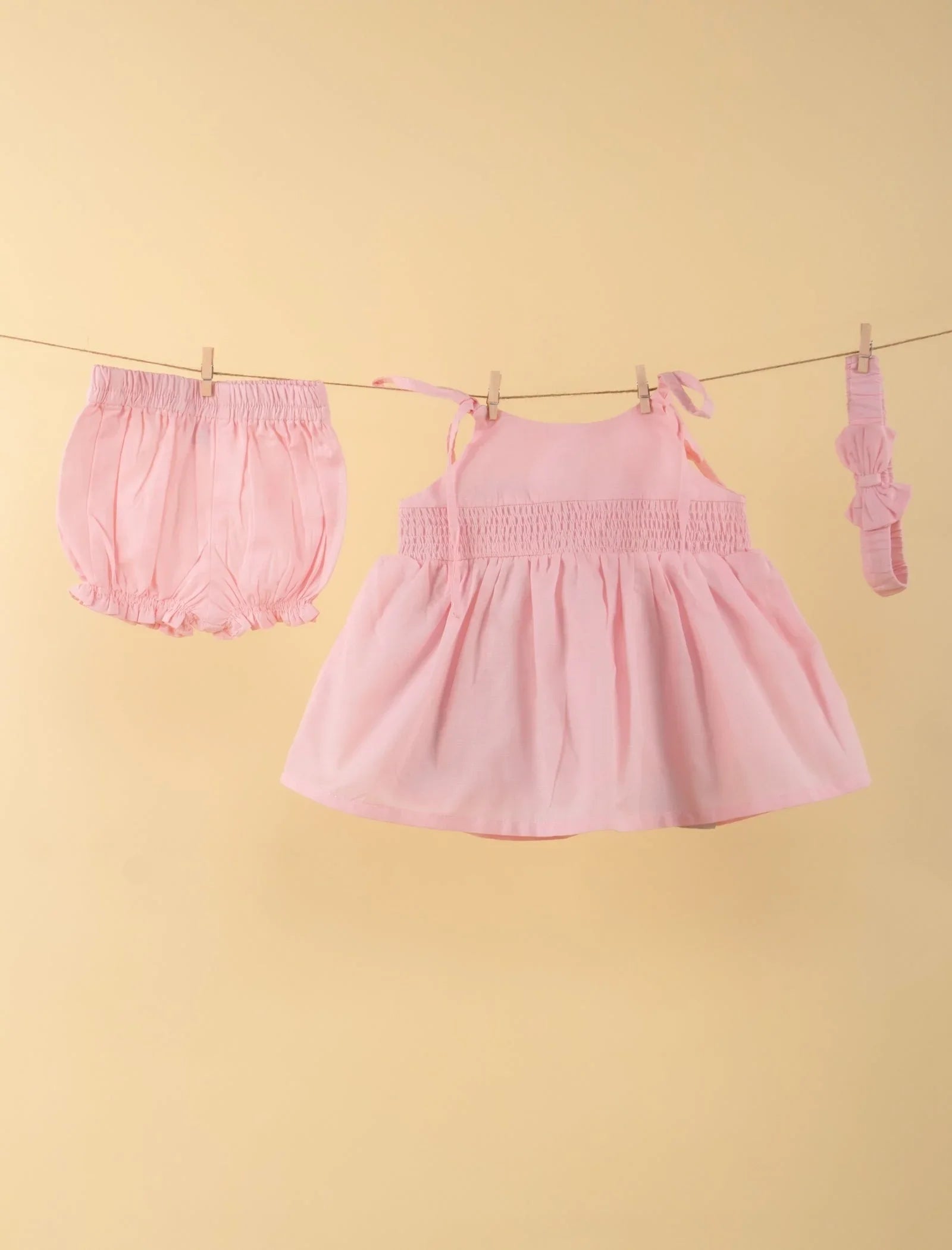 Pink baby girl dress set with bloomers and headband, hung on a clothesline, beige background
