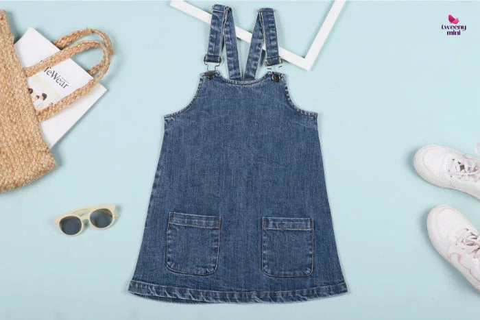 Girls' denim dungaree dress with front pockets, flat lay with shoes and accessories