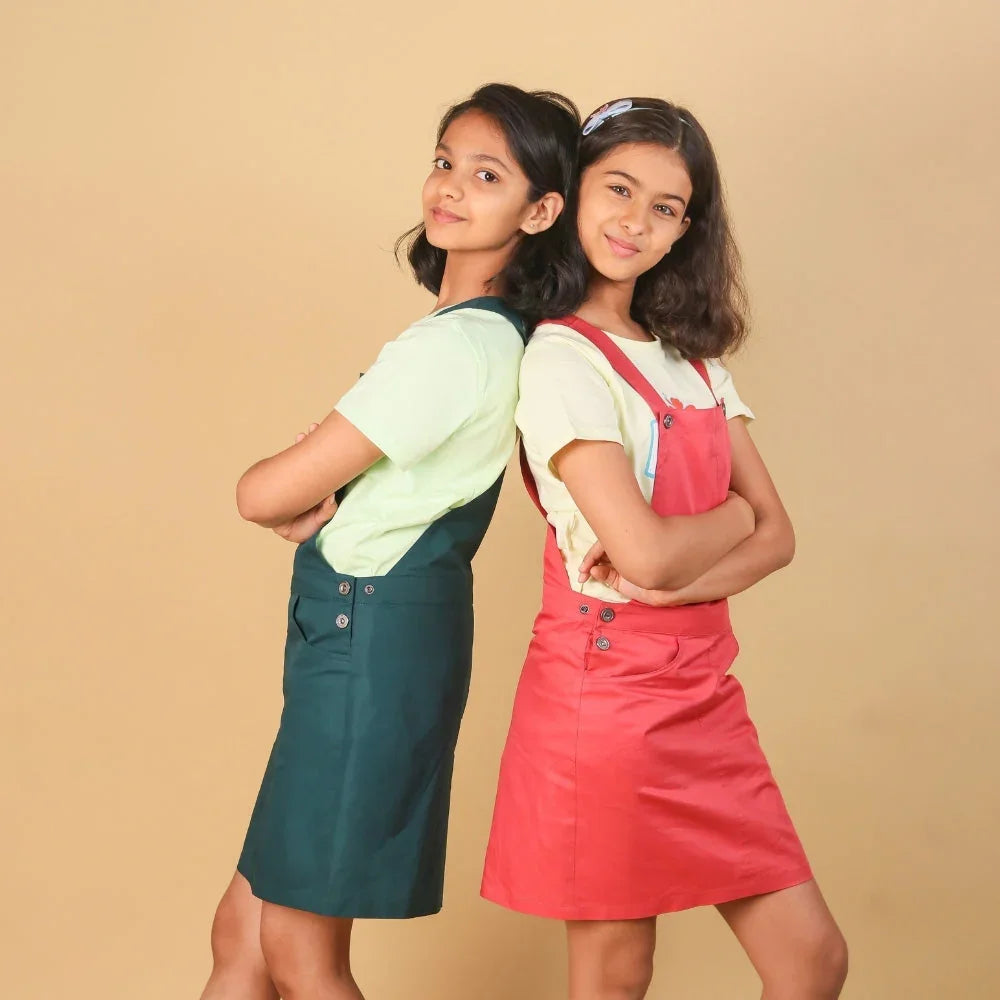 Two girls in casual dungaree dresses, one teal and one pink, posing back to back on beige background.