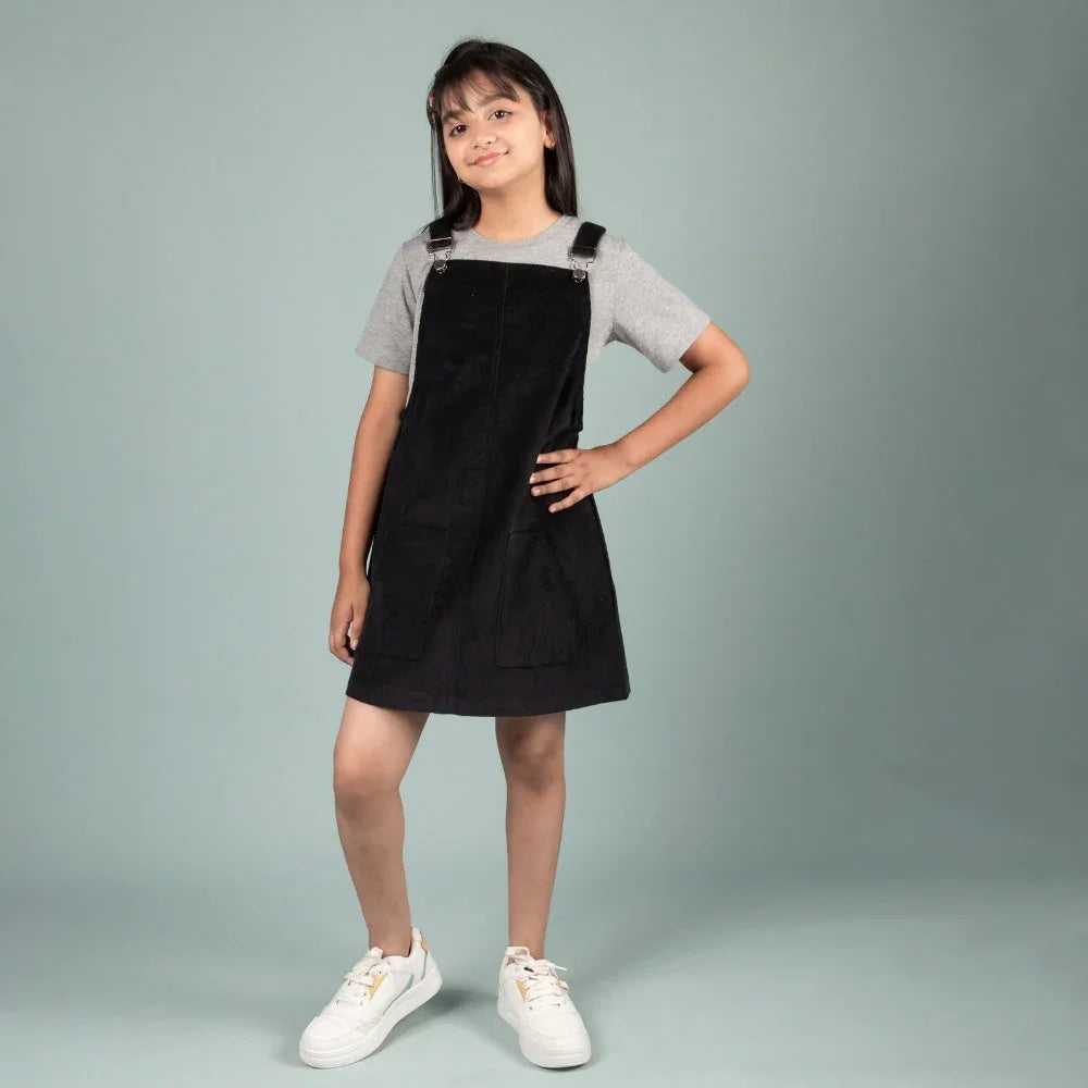 Girl in black pinafore dress and grey t-shirt, posing against a plain background