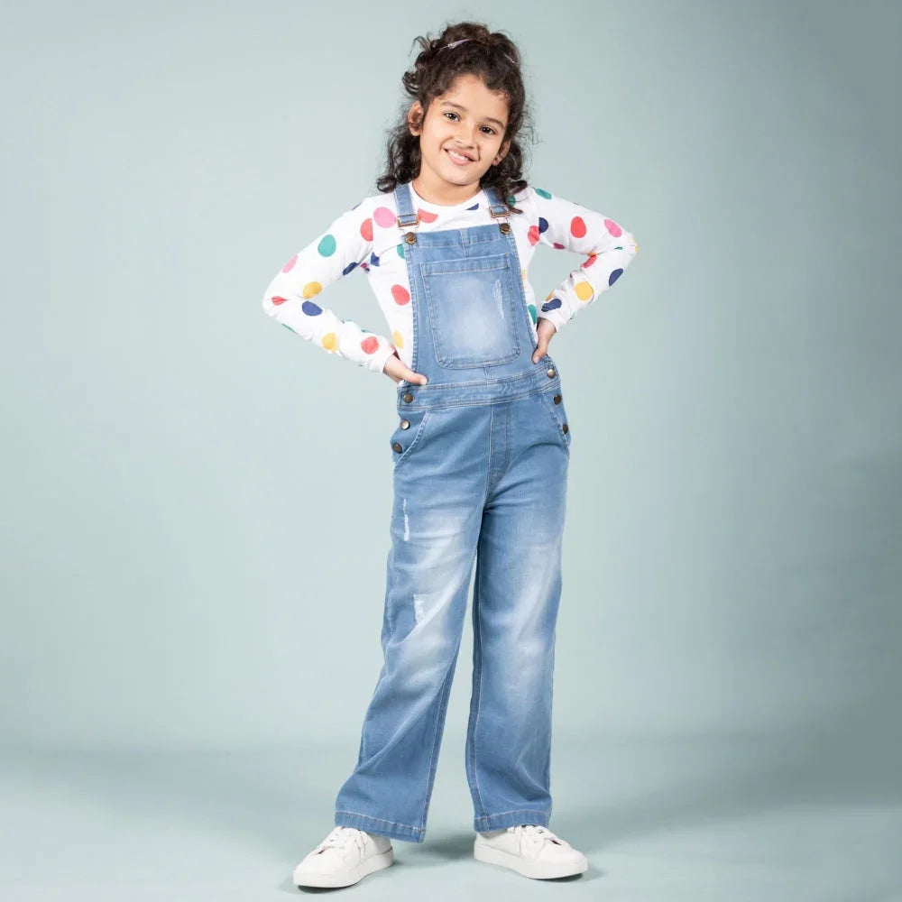 Girl in denim dungarees and colorful polka dot top posing against plain background