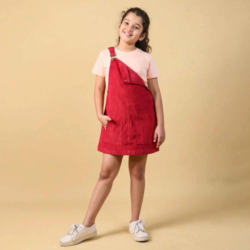 Smiling girl in a pink t-shirt and red dungaree dress, posing against a beige background.