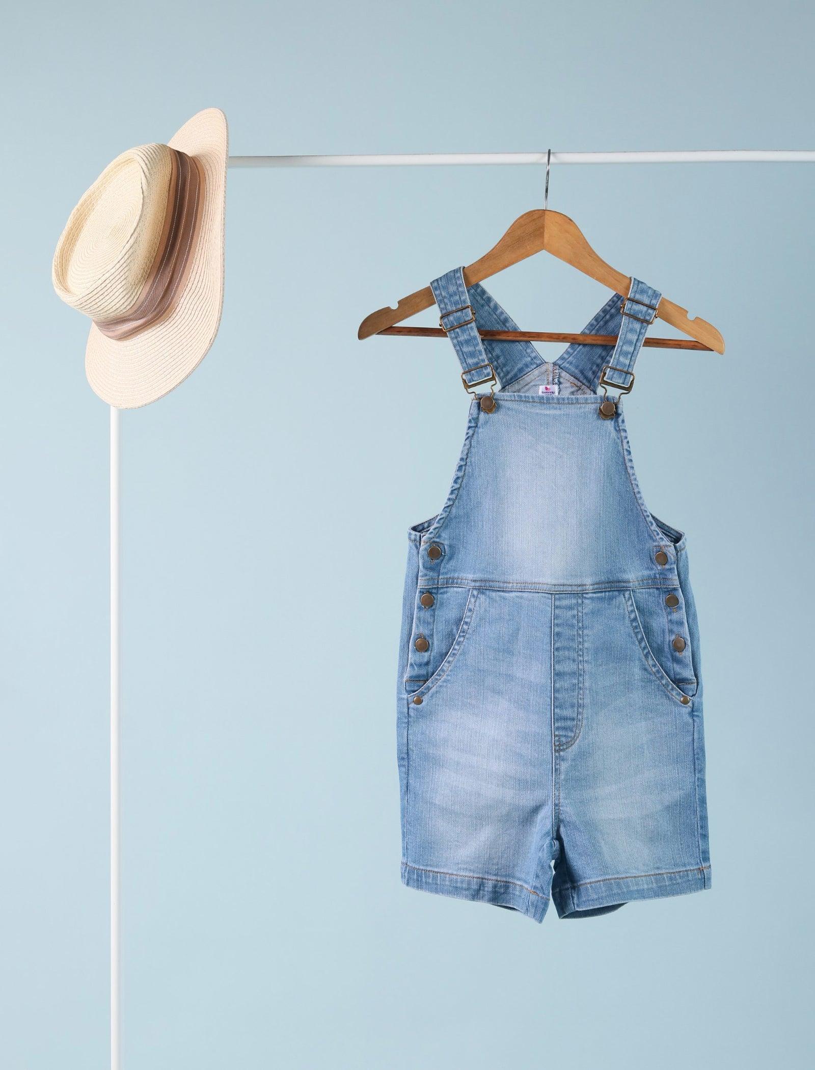 Light blue denim dungarees for girls aged 4, hanging on a wooden hanger near a straw hat