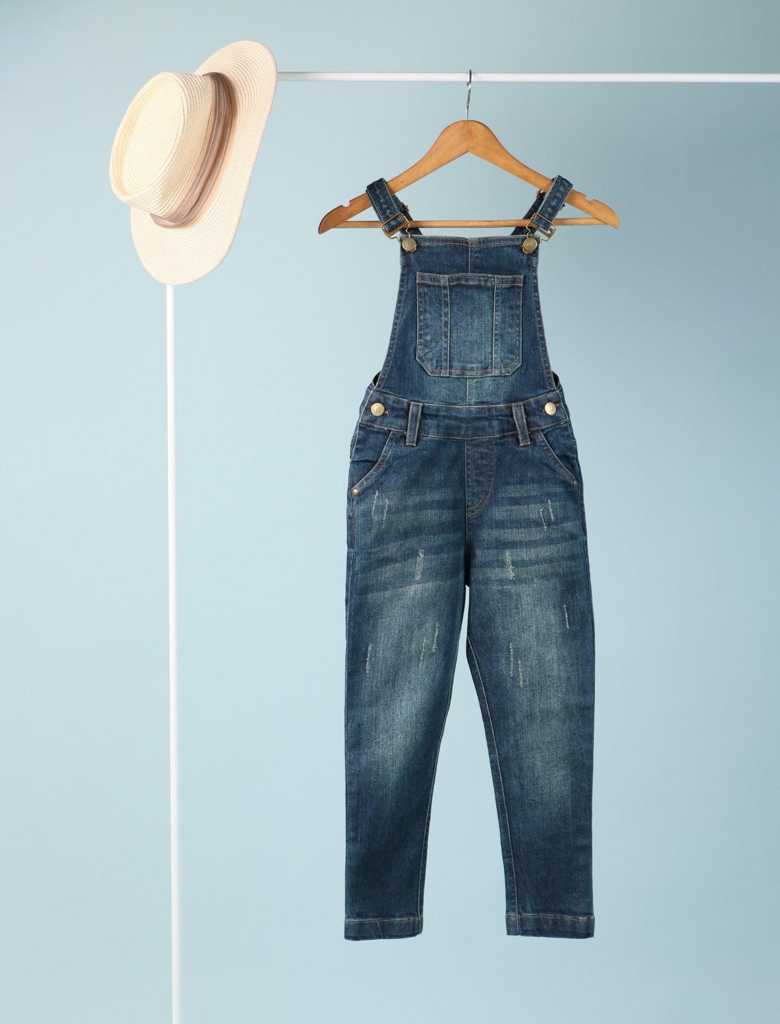Denim dungarees for girls age 6 with front pocket, hanging on a wooden hanger next to a hat
