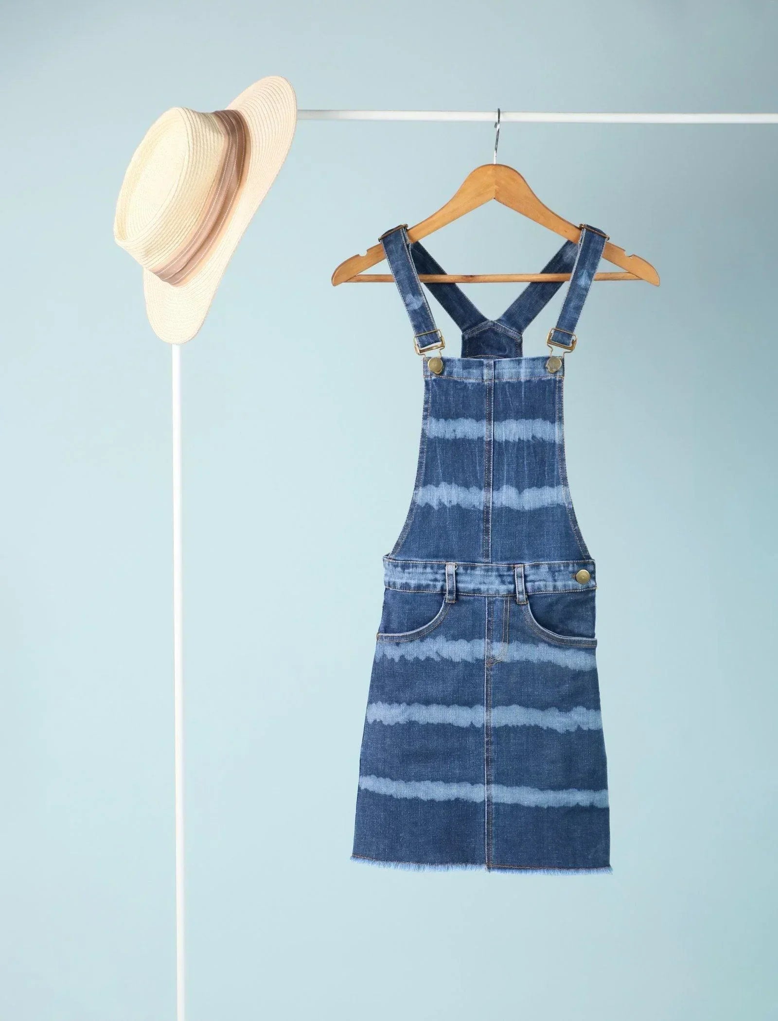 Denim dungaree dress for girls with blue tie-dye stripes, hanging on wooden hanger, straw hat nearby