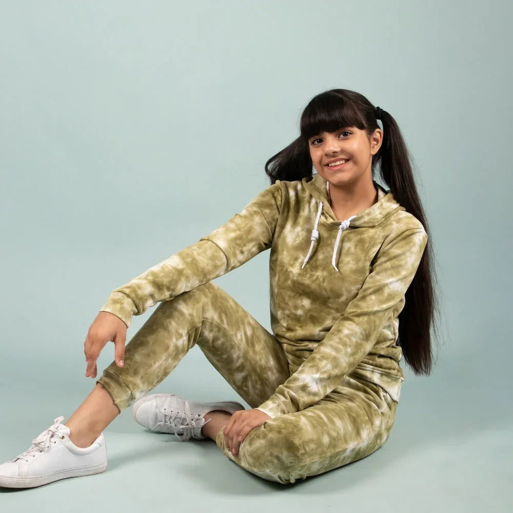 Smiling girl in green tie-dye jogger set and white sneakers sitting on light blue background