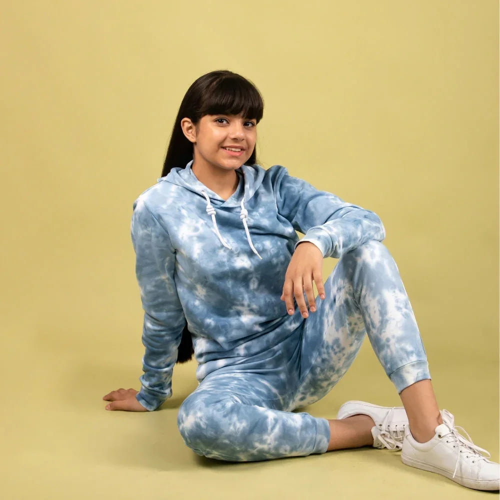 Girl in blue tie-dye jogger set and white sneakers sitting against yellow background