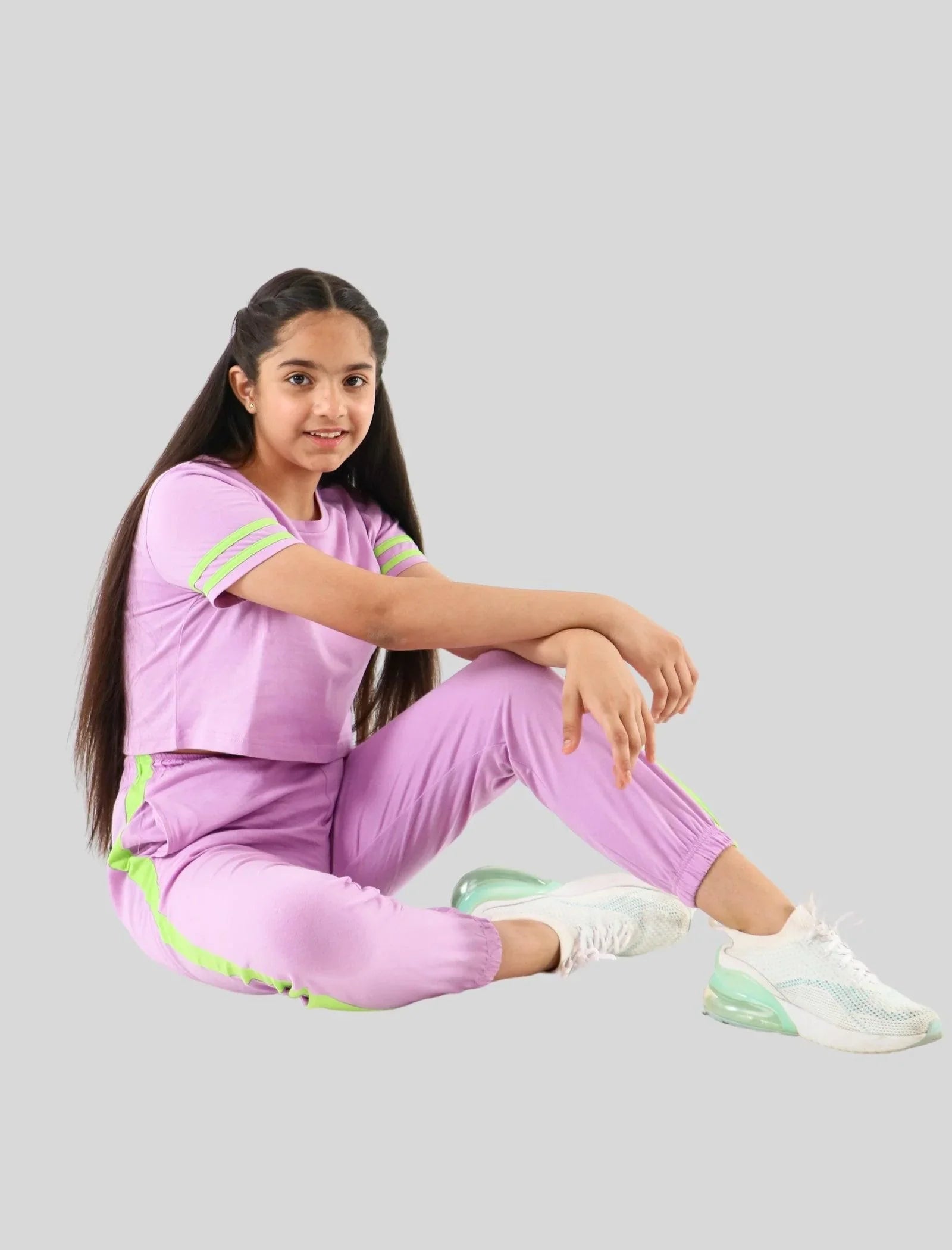 Teen girl in lilac jogger set with green stripes, sitting in white sneakers on a white background