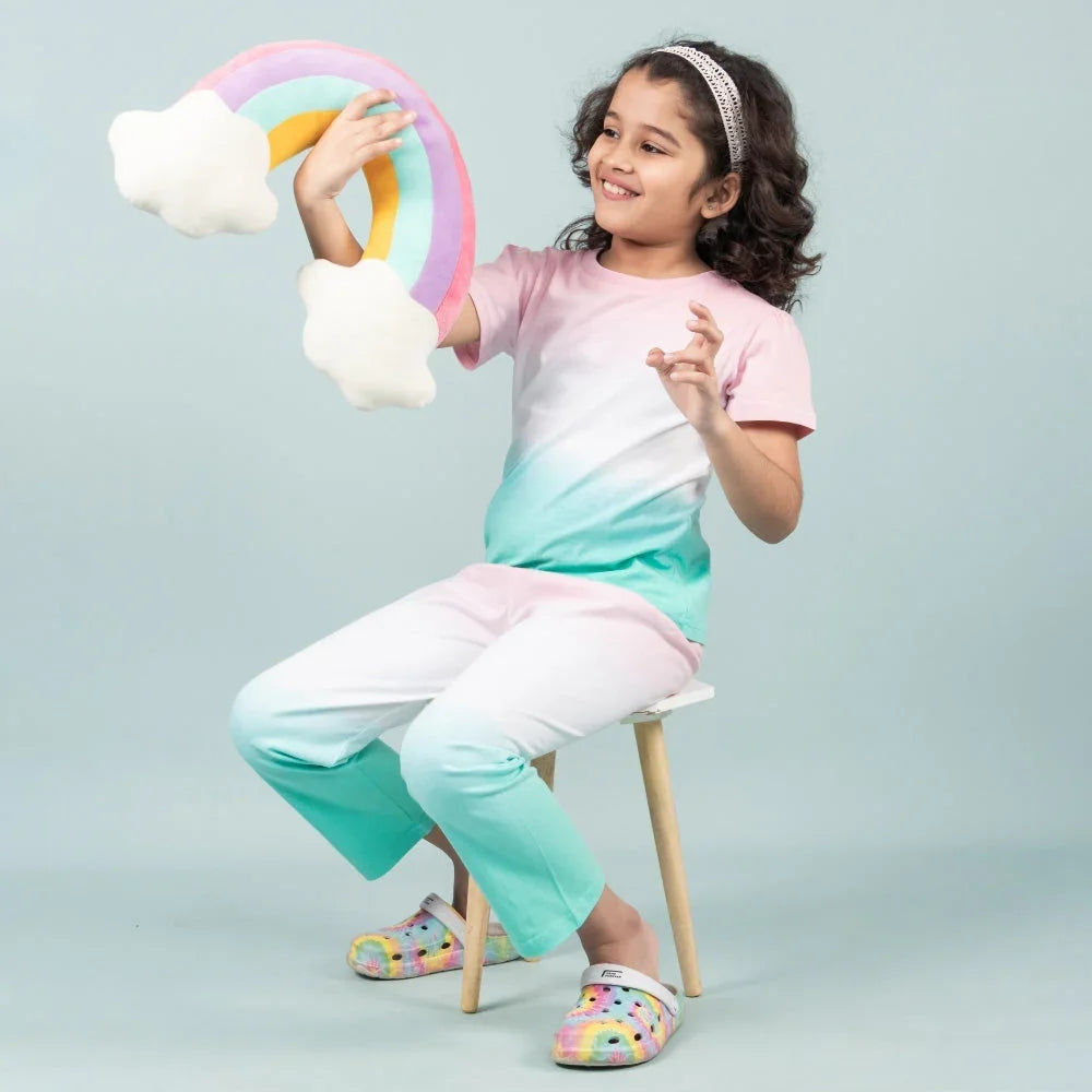 Young girl in pastel night suit for girls, holding rainbow pillow, sitting on stool
