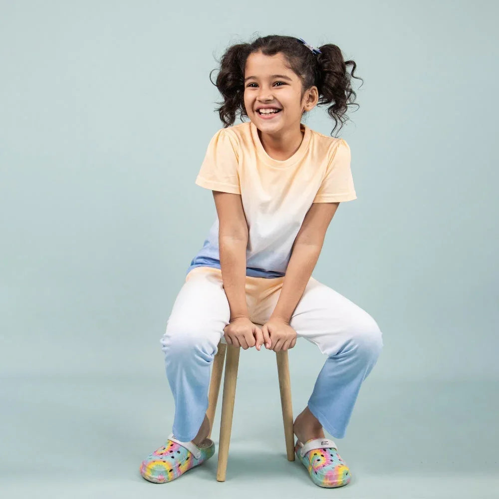Smiling girl in pastel night suit and colorful slippers sitting on a stool, kids wear fashion.
