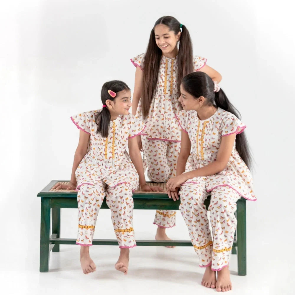 Three girls in matching printed nightdresses with flutter sleeves and pink trim, sitting together on a bench in a studio setting.