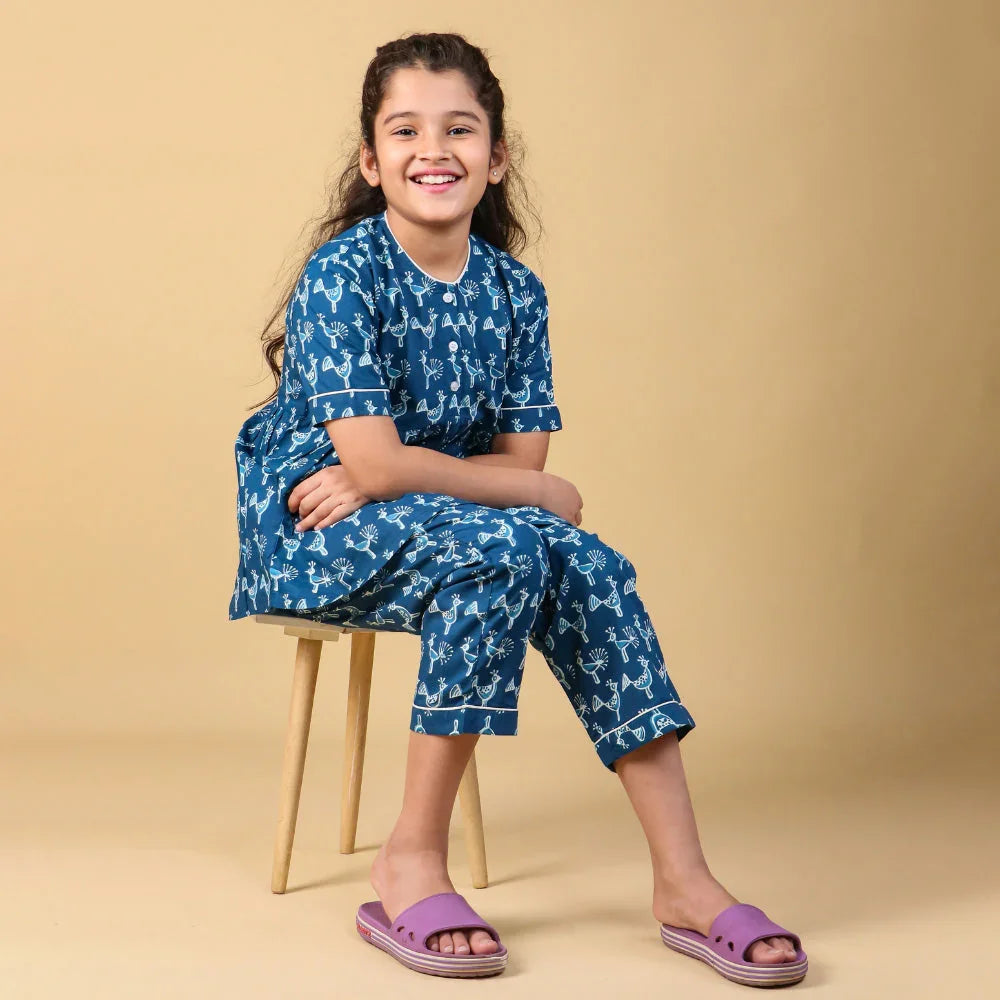 Smiling girl in blue printed night suit and purple slippers sitting on a wooden stool