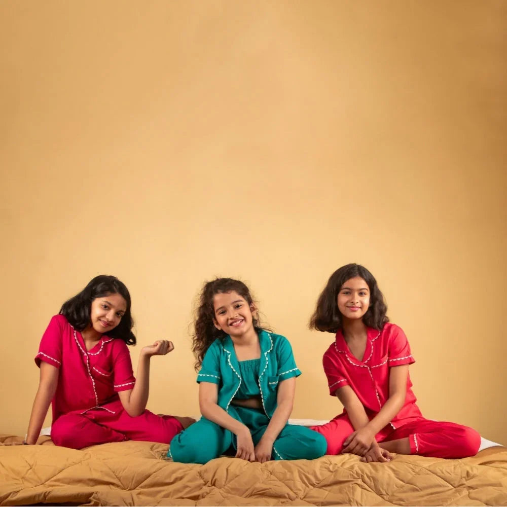 Three girls aged around 8 wearing colorful nightwear sets, sitting on a bed against a beige background.