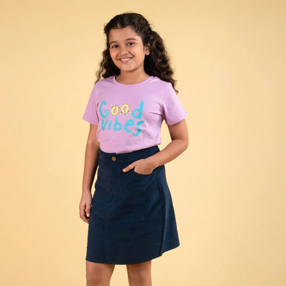 Smiling girl wearing purple graphic t-shirt and navy blue skirt, hands in pockets, beige background.