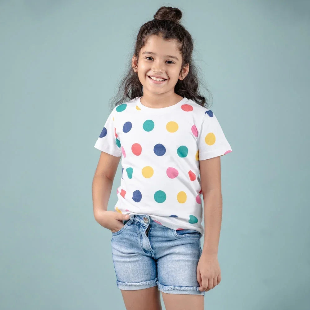 Smiling girl wearing a white polka dot t-shirt and denim shorts, standing against a light blue background