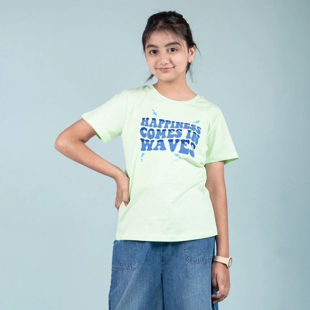 Girl wearing light green graphic t-shirt with 'Happiness Comes in Waves' text and denim shorts