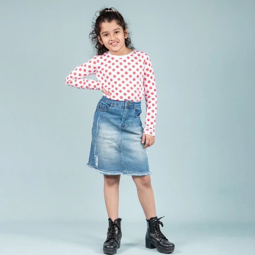 Smiling girl wearing white red polka dot top, denim skirt, and black boots on light background