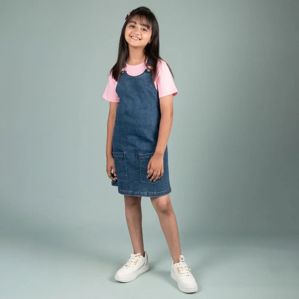 Smiling girl wearing a denim pinafore dress over a pink t-shirt and white sneakers