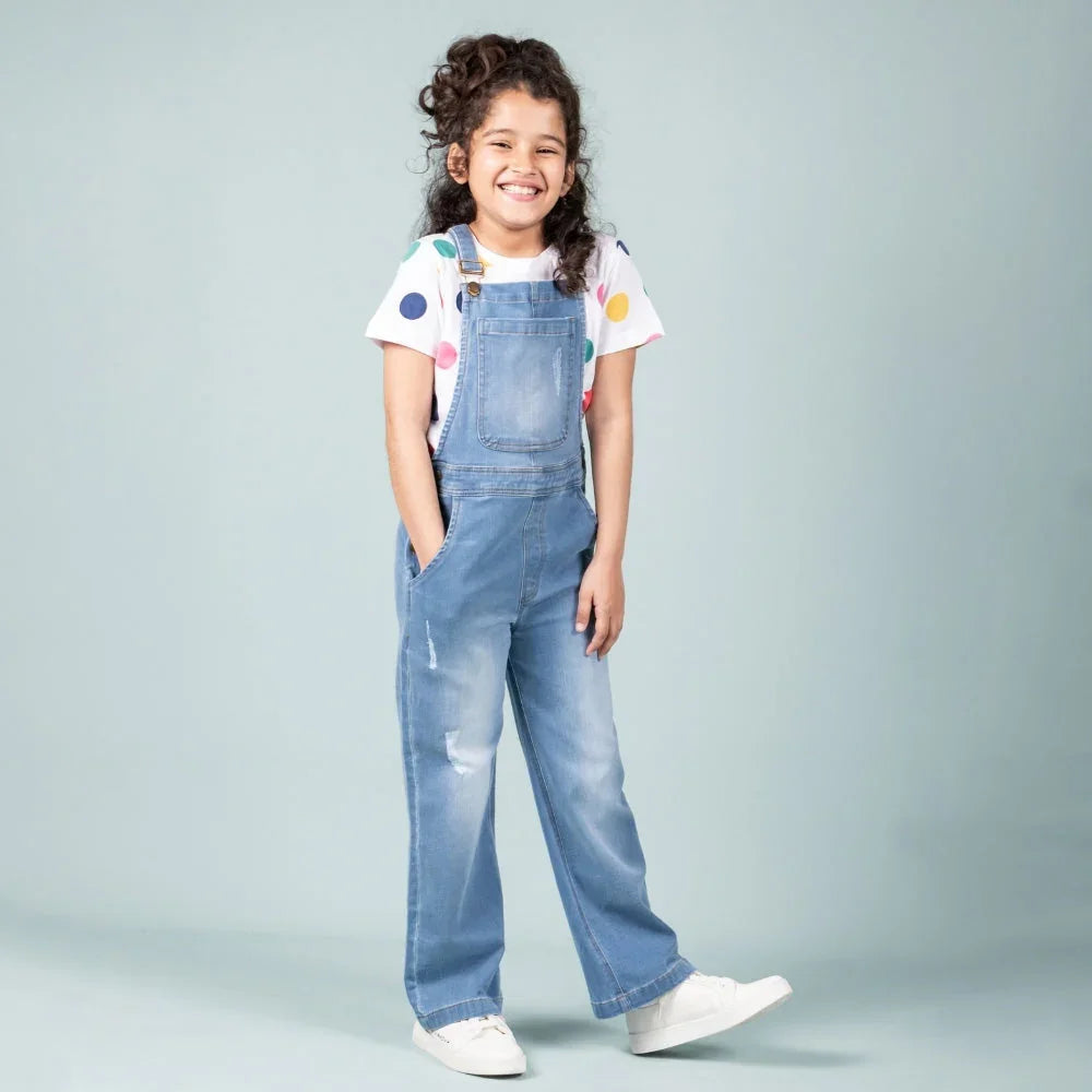 Smiling girl wearing light blue denim dungarees, white polka dot t-shirt, and white sneakers.